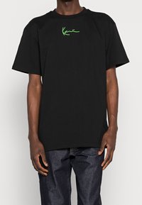 T-shirt in cotone nero con una vestibilità ampia, caratterizzata da maniche corte e un logo verde distintivo sul petto. Colore uniforme, texture liscia.