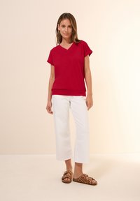 Haut rouge à manches courtes en V, fabriqué en tissu doux, associé à un pantalon blanc cropped et des sandales marron à pois blancs.