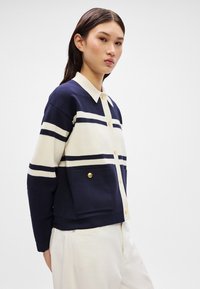 Jūrinės ir grietinėlės spalvų juostuota striukė su apykakle, mygtukų uždarymu ir priekinėmis kišenėmis, kurių akcentas - auksinis mygtukas. Minkšta audinio tekstūra.