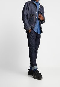 Dark denim jacket with two chest pockets, layered over a light blue button-up shirt; paired with dark pants and black shoes.