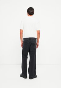 Mand står med ryggen til, iført en hvid kortærmet skjorte, sorte løse jeans og sorte sko med kraftig sål foran en lys hvid baggrund.