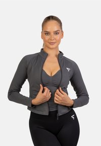 Ľahká, priliehavá športová bunda v tmavošedej farbe s vysokým golierom a zipsom. Má dlhé rukávy a malú logovú akcentáciu na hrudi.