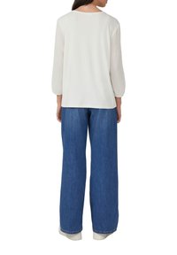 Vrouw in een witte top met lange mouwen en gestructureerde mouwen, met een blauwe wijde jeans, staande met haar rug naar de camera op een witte achtergrond.