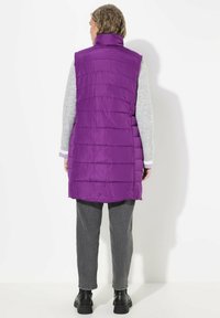 Langer lila Puffer-Weste mit hohem Kragen und gestepptem Design. Getragen über einem grauen Langarmoberteil, kombiniert mit dunklen Hosen und schwarzen Stiefeln.