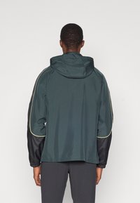 Donkergroene sportjacket voor heren met capuchon, met zwarte mouwen, elastische manchetten en reflecterende gele accenten langs de schoudernaden.