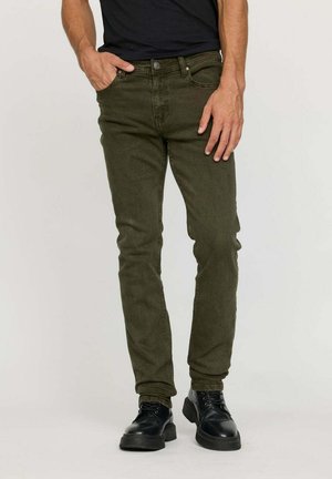 Homme portant un jean slim vert olive avec des chaussures noires en cuir lacées, debout devant un fond neutre.