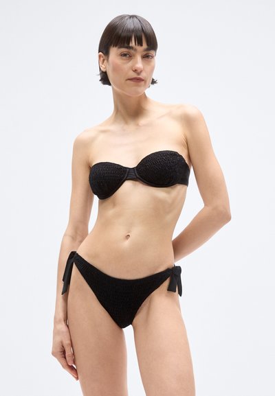 Črni bikini komplet z zgornjim delom bandeau z teksturo in spodnjim delom z zavezovanjem na straneh. Obe kosu imata grdo vzorec in gladko tkaninsko obdelavo.