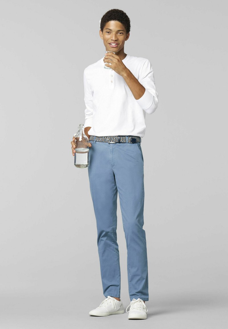 Maglietta henley bianca a maniche lunghe, chinos azzurri chiari e sneakers bianche. Il modello tiene in mano un bicchiere d'acqua e una bottiglia, in piedi contro uno sfondo grigio.