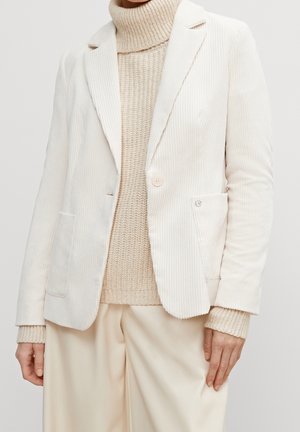 Kobieta nosi kremową sztruksową marynarkę na beżowym dzianym swetrze z golfem i pasujących kremowych szerokich spodniach.