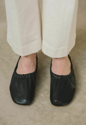 Zapatos negros de cuero sin cordones con punta redonda, parte superior elástica y suela plana, combinados con pantalones de pierna ancha color crema. Textura suave y diseño minimalista.