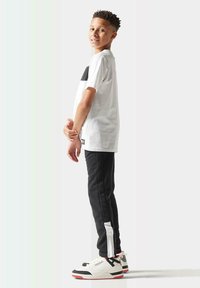 Camiseta blanca con panel negro, pantalones negros con rayas laterales blancas y zapatillas blancas con acentos rojos. Pose de pie con las manos entrelazadas.