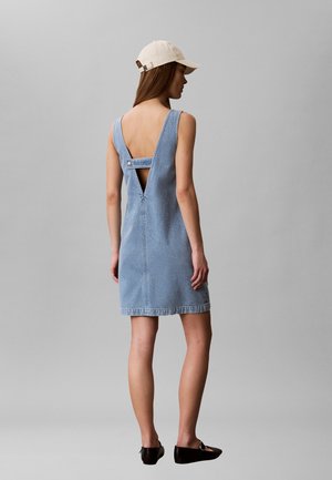 Kvinde iført en lyseblå denimærmeløs kjole med dyb V-udskæring bagpå og en beige baseballkasket, stående foran en ensfarvet grå baggrund.