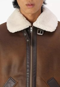 Giacca di pelle marrone con colletto bianco soffice, dotata di una zip frontale, hardware argentato e una finitura testurizzata. I dettagli includono rifiniture in pelle scura.