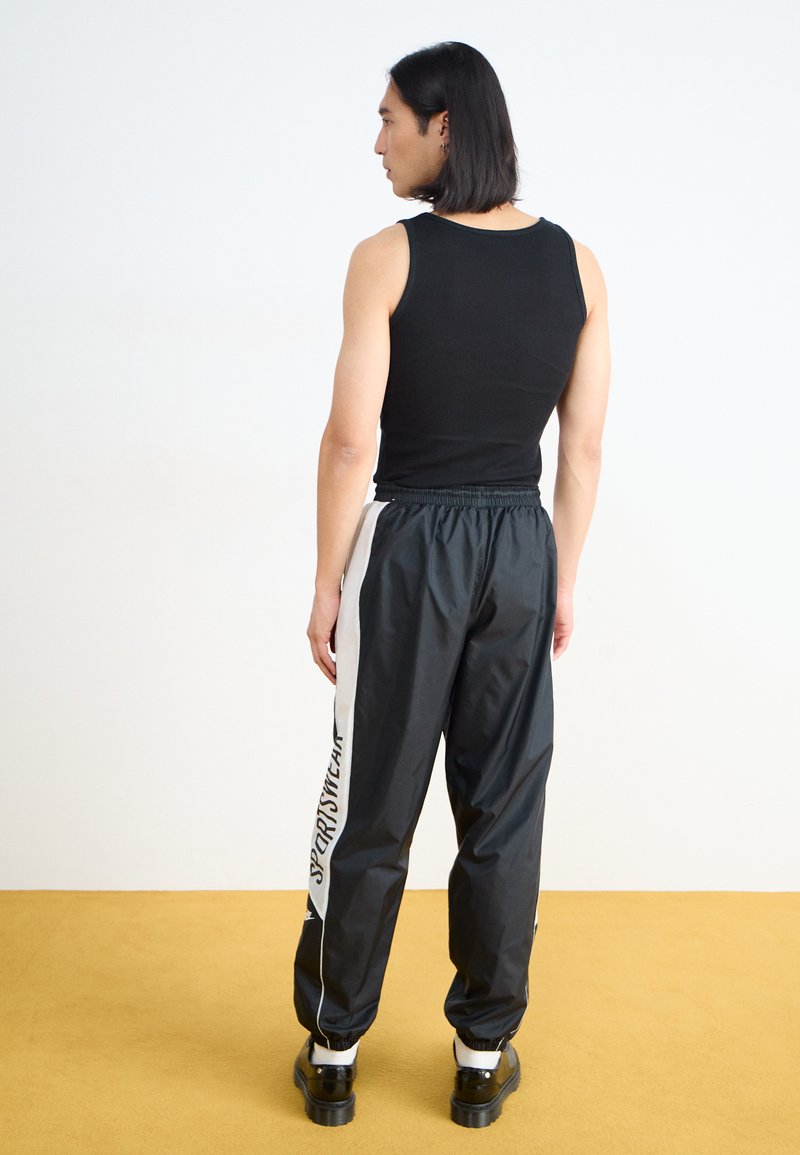 Débardeur noir à côtes associé à un pantalon de jogging noir à taille élastique, avec une bande latérale blanche et du texte. Chaussures à semelle épaisse noires.