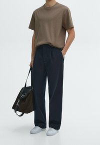 Man wearing brown short-sleeve shirt, navy trousers, and white sneakers holding a brown and black shoulder bag on white background.