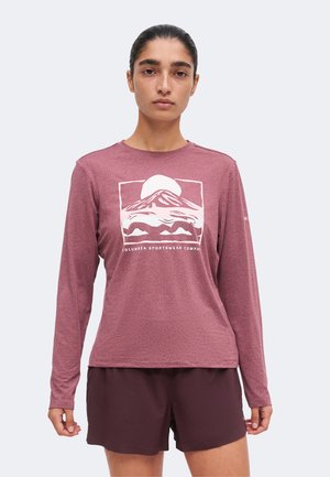 Kvinde iført langærmet vinrød skjorte med bjergmotiv og shorts, stående mod en ensfarvet lys baggrund, med front mod kameraet.