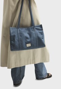 Borsa in denim con doppi manici, caratterizzata da una forma rettangolare, colore azzurro chiaro e una piccola etichetta sul davanti.