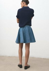 Femme debout, de dos, portant un haut à manches courtes bleu marine, une jupe en denim évasée bleue et des chaussures plates noires sur un fond neutre.