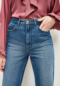 Jeans de mezclilla azul de talle alto con cierre de botón, cinco bolsillos y detalles de costura en color naranja, combinados con una blusa rosa.