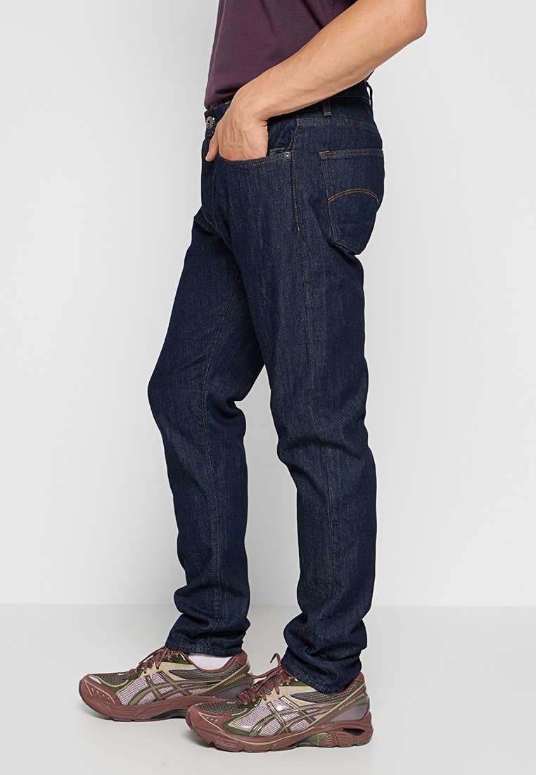 Persona che indossa jeans blu scuro e sneakers marrone-verdi, in piedi con una mano in tasca contro uno sfondo chiaro e uniforme.