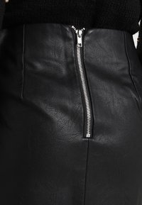 Černá sukně z umělé kůže se lesklým stříbrným zipem probíhajícím vertikálně středem vpředu, spárovaná s černým pleteným topem.