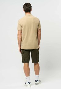 Muž stojící zády, oblečený v béžovém polokošili s krátkým rukávem, tmavě zelených cargo šortkách, bílých ponožkách a bílých teniskách s modrými a červenými akcenty.