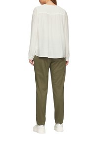 Blouse blanc texturé à manches longues, associé à un pantalon vert olive. Le modèle porte des baskets blanches épaisses. Vue de dos ; aucun détail saillant visible.