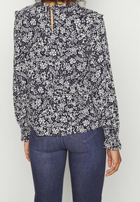 Blusa blu navy con motivo floreale bianco, con maniche lunghe e polsini arricciati, e dettagli sulla schiena a forma di chiave. Tessuto morbido con finitura texturizzata.