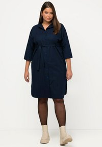 Vestito camicia blu navy con colletto, chiusura a bottoni sul davanti, cintura da annodare in vita, maniche a metà lunghezza e spacchetti laterali. Tessuto morbido.
