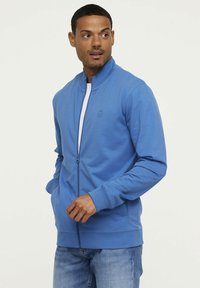 Sweatshirt zippé bleu avec un col montant, en tissu léger, poches latérales, poignets côtelés et un logo discret sur la poitrine.