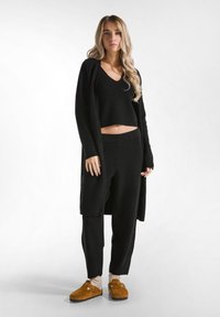 Cardigan nero a maglia, top corto e pantaloni aderenti; pantofole marroni pelose con cinghie regolabili; texture morbide e lisce.