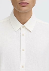 Camisa de color beige claro con cuello, hecha de tejido de fibra, que presenta un cierre frontal con botones, textura visible y diseño de cuello estándar.