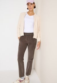 Licht beige bomberjack, witte t-shirt, donkerbruine broek met trekkoord en crèmekleurige sneakers met bordeaux accenten. Gladde texturen overal.