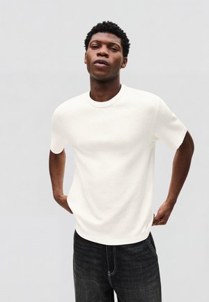 Ung mand med kort krøllet hår iført en hvid, kortærmet trøje og mørke jeans foran en lys grå baggrund.