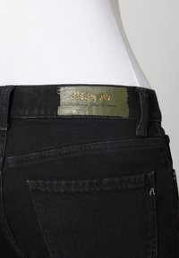 Jeans de mezclilla negros con un parche de cuero verde que presenta la marca en dorado "REPLAY" en la cintura, y detalles de costura sutiles en los bolsillos traseros.