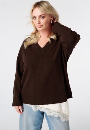 Blonde vrouw met een donkerbruine oversized V-hals trui over een witte top met kant en lichtblauwe spijkerbroek, poserend met één hand bij het haar.