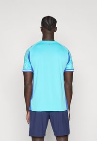 Αθλητική μπλούζα ανοιχτό μπλε με κοντά μανίκια, με σχέδια στα μανίκια και μπλε λεπτομέρειες. Συνδυάζεται με σκούρο μπλε σορτς.