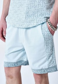 Shorts de sport bleu clair en tissu doux, arborant un motif géométrique avec la marque "PROJECT X PARIS" le long de la taille et sur le côté.