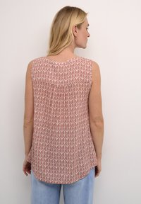 Blouse sans manches, rouge à motif avec un design de feuilles, détails rassemblés au dos et ourlet arrondi. Portée avec un jean bleu clair.