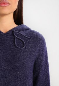 Sudadera de punto morada con detalle de cordón en el escote. Textura suave y corte holgado, que muestra un diseño acogedor.