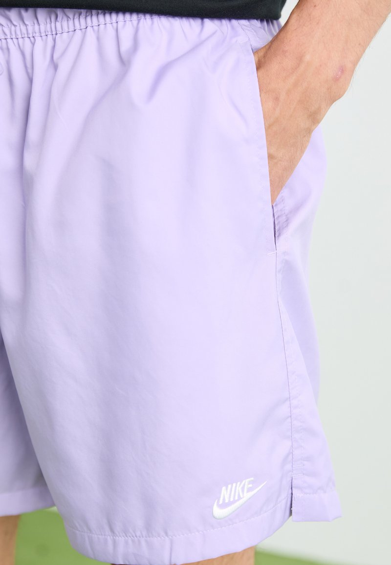 Shorts Nike lilas clairs avec une taille élastique, des poches latérales et un logo blanc en bas de l'ourlet. Texture de tissu lisse.
