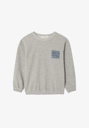 Usmievavé dieťa s kučeravými vlasmi oblečené v sivej mikine, modrých šortkách a béžových slip-on topánkach stojace pred jednofarebným pozadím.