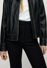 Veste en cuir noir avec fermeture éclair à l'avant, présentant une texture lisse et des coutures contrastées. Associée à un pantalon noir taille haute.