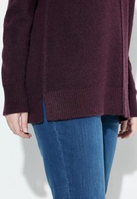 Maglione in maglia bordeaux con polsini e orlo a coste, caratterizzato da aperture laterali. Abbinato a jeans in denim blu, che mostrano uno stile casual.