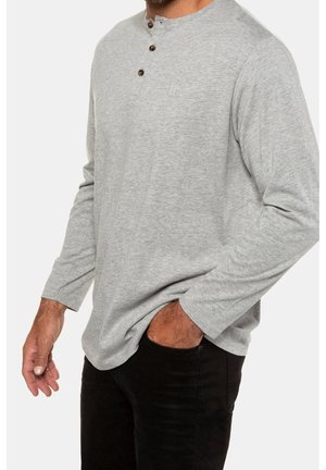 Homme portant un t-shirt henley gris clair à manches longues avec des boutons, main gauche dans la poche du pantalon noir, bras droit détendu le long du corps.