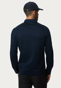 Marineblaue Langarmbluse mit kragenförmigem Design, lockerer Passform und Knopfmanschetten. Getragen mit einer passenden Mütze, die eine glatte Textur zeigt.