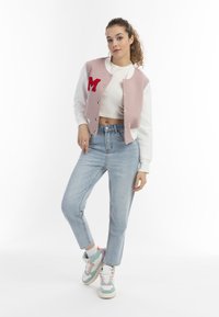 Rosa und weiße, kurz geschnittene Collegejacke mit "M"-Patch, kombiniert mit hellblauen, hochtaillierten Jeans und mehrfarbigen Sneakers.