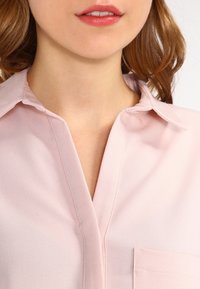 Jasnoróżowa bluzka z kołnierzykiem, z dekoltem w kształcie litery V i kieszonką po lewej stronie. Tkanina wydaje się gładka i lekka.