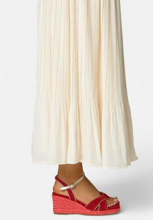 Pieds chaussés de sandales compensées rouges avec des brides dorées à la cheville, sous une longue jupe plissée crème, présentés sur un fond clair uni.