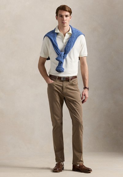 Lyseblå genser knyttet over en hvit polotrøye, kombinert med beige bukser og brune sko. Enkel, klassisk design og nøytralt fargevalg.